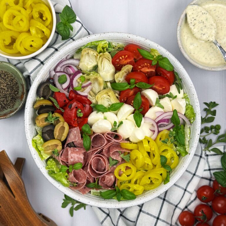 A beautiful white bowl, full of banana peppers, tomatoes onions and more with a side of creamy italian dressing.