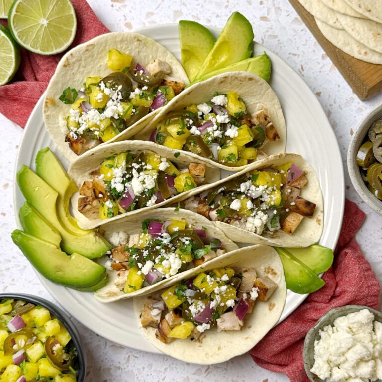 A white plate with six BBq Grilled Chicken Tacos on its. Sliced Avocado around the tacos on the plate and a bowl of Pineapple Salsa on the side.