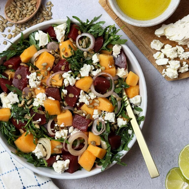 Sweet Beet and Melon Salad in a white bowl on a counter with dressing and toppings on a wooden board.