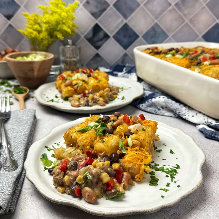 Two round white plates with Tater Tot Casserole with Cowboy Caviar and Ground Beef on them with the full pan of Casserole next to it.