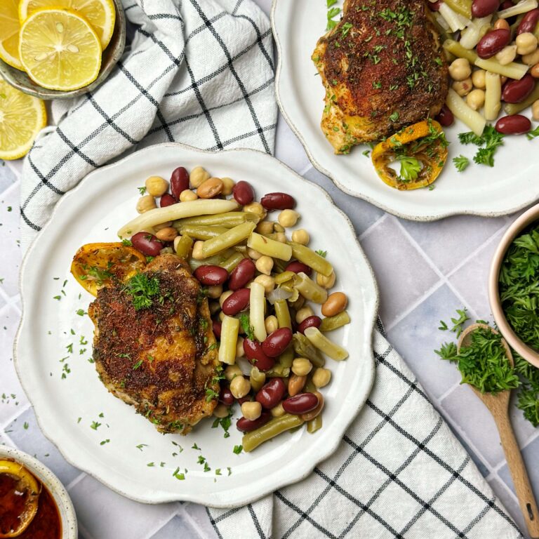 A white dinner plate with Seasoned Chicken Thighs served with a side of Five Bean Salad.